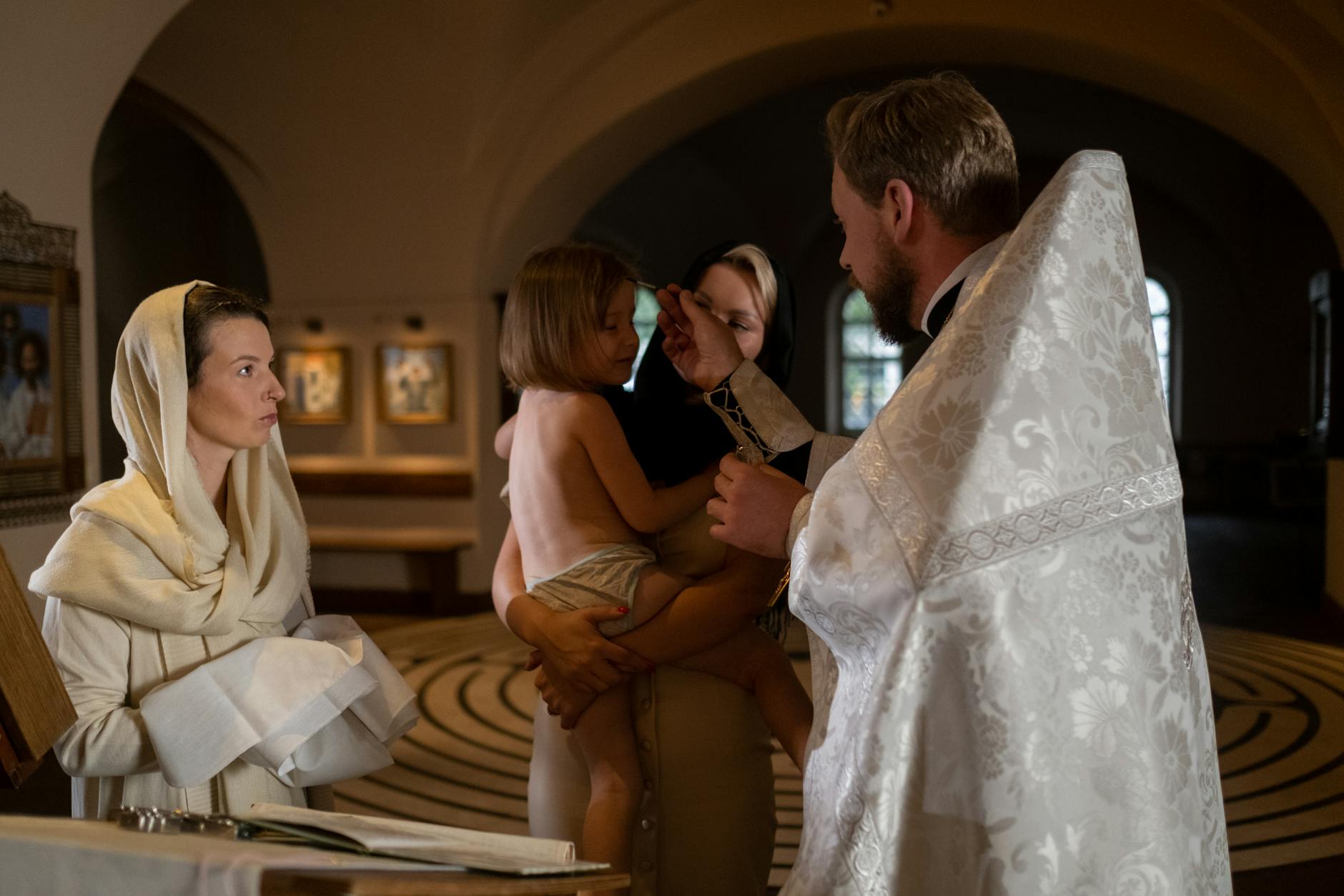 Kapłan chrzci niemowlę w kościele, obok stoją członkowie rodziny