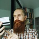 Brodaty mężczyzna sprawdza firmę w telefonie, trzymając kartę płatniczą