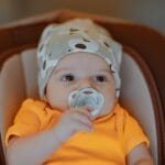 Jak przygotować wyprawkę dla noworodka na pierwszą podróż samochodem Niemowlę w pomarańczowym body z czapką i smoczkiem w domu