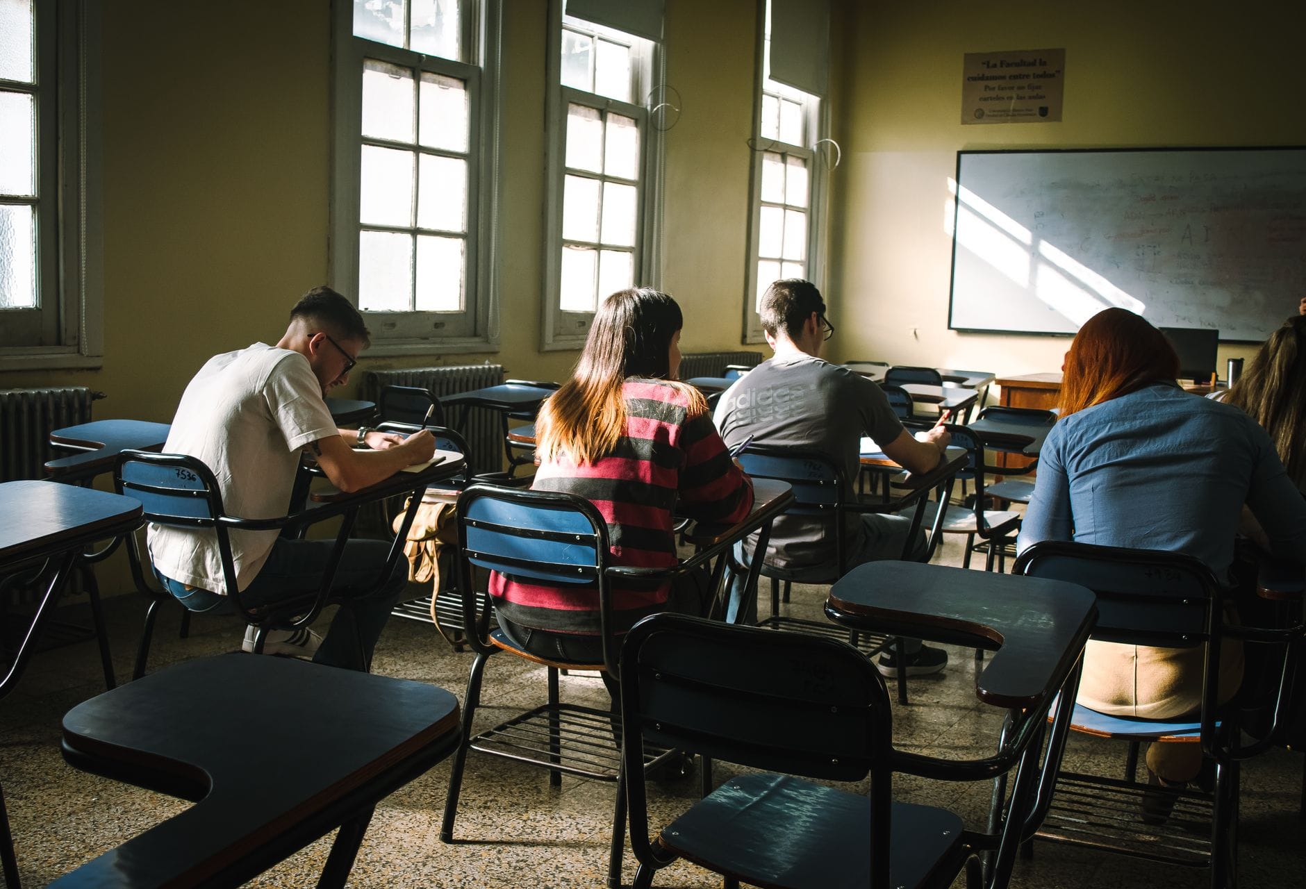 Uczniowie piszący test w słonecznej sali pełnej ławek i okien